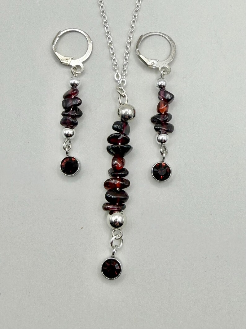 May include: A jewelry set featuring a necklace and a pair of earrings. The necklace has a silver chain with a pendant of stacked, dark red gemstones and silver beads, and a single red gemstone at the bottom. The earrings have a similar design.