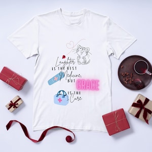 Puede incluir: Camiseta blanca con un diseño gráfico que presenta un gato de dibujos animados, una venda y un bolso médico. El texto dice: "La risa es la mejor medicina, pero la gracia es la cura."