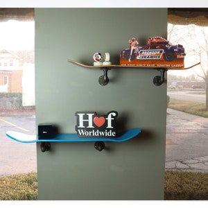 May include: Two skateboard shelves mounted on a wall. The top shelf is made of wood and has a sticker that says "Bronson Ceramics" and "If you ain't fast you're last!" The bottom shelf is blue and has a black and white sticker that says "Hof Worldwide" with a red heart.