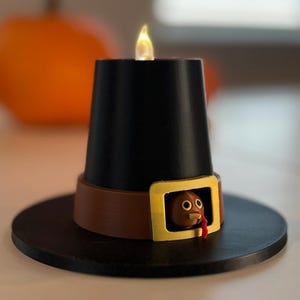 May include: A black candle holder shaped like a pilgrim hat with a brown band and a gold buckle. A small turkey is visible in the buckle. The candle has a flickering flame. Two orange pumpkins are in the background.
