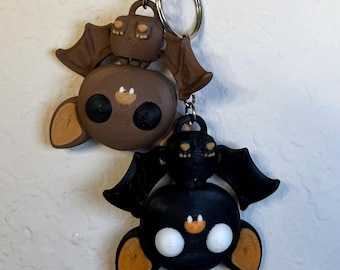 May include: A keychain featuring two bat-shaped charms. The top charm is brown with tan wings and a small bag. The bottom charm is black with tan accents. Both charms have large black eyes and are attached to a silver keyring.