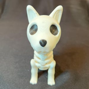 Könnte beinhalten: Eine weiße Hundefigur mit Skelett-Design. Der Hund hat große, dunkle Augenhöhlen und eine schwarze Nase. Die Figur steht auf allen Vieren, wobei Rippen und Knochen der Beine sichtbar sind.