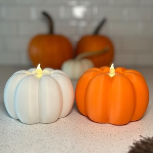 May include: Two decorative pumpkin-shaped lights, one white and one orange, with a flickering candle-like glow. The pumpkins are on a countertop, with larger pumpkins in the background, creating a fall or Halloween theme.