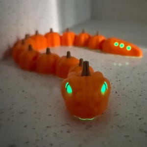 May include: An orange pumpkin snake with glowing green eyes and accents. The snake is made of individual pumpkins with brown stems. The eyes and three circles on the tail glow green. A unique Halloween decoration.