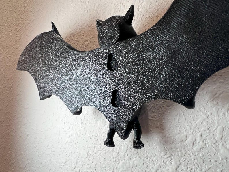 Op de afbeelding: Een zwarte vleermuisvormige wanddecoratie met getextureerde vleugels en een gedetailleerd lichaam. De vleermuis heeft twee gaten om op te hangen en is gemaakt van een duurzaam materiaal. De vleugels zijn wijd gespreid en de vleermuis is op een witte muur gemonteerd.