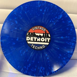 Op de afbeelding: Een blauwe vinylplaat met witte spetters. Het platenlabel heeft de tekst "Vintage Detroit Techno" met een rood-wit-blauw kleurenschema en een silhouet van de skyline van Detroit.