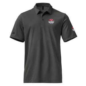 Peut inclure: Un polo à manches courtes gris foncé avec un logo Adidas noir sur la manche gauche et un logo circulaire blanc sur la manche droite. Le logo indique "Vintage Detroit Techno" en texte blanc.