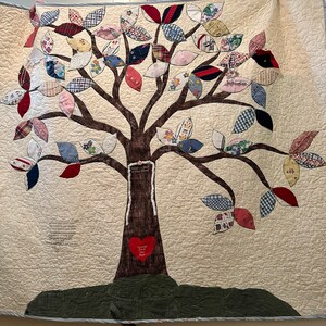 May include: A brown fabric quilt with a tree design. The tree is made of fabric scraps in various colors and patterns. The leaves are shaped like hearts and are attached to the branches. The tree trunk is brown and has a heart-shaped patch sewn on it. The quilt is surrounded by a white border.