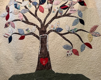 May include: A brown fabric quilt with a tree design. The tree is made of fabric scraps in various colors and patterns. The leaves are shaped like hearts and are attached to the branches. The tree trunk is brown and has a heart-shaped patch sewn on it. The quilt is surrounded by a white border.