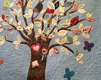 May include: A blue quilted fabric tree with brown branches and leaves made from different fabrics. The leaves have various words and phrases on them, including "Be Kind", "Best Ever", and "I am a Skier". There are also butterflies and a rainbow on the fabric.