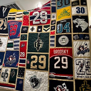 May include: A handmade quilt featuring patches of sports jerseys and team logos. The quilt showcases various colors, including red, black, green, and blue, with numbers and team names prominently displayed. The quilt is a patchwork design, with each square containing a different jersey or logo.