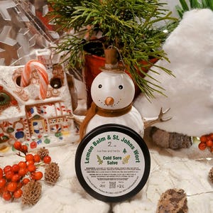 Può includere: Una scatola di latta nera di balsamo al limone e erba di San Giovanni per il trattamento delle labbra screpolate, con un'etichetta verde e testo bianco. L'etichetta dice "2 oz live free and herbs". La scatola è appoggiata su una superficie bianca con un pupazzo di neve e una casetta di pan di zenzero sullo sfondo.