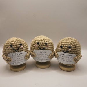 May include: Three crocheted potato plushies, each holding a sign with a positive message. The signs read: "Positive Potato I may be a tiny Potato, but I Believe in you! Go do your thing!"  "Positive Potato Small potatoes Can still make a Big impact! Keep Pushing forward!"  "Positive Potato Life was all out of Lemons, so here is A potato instead!"