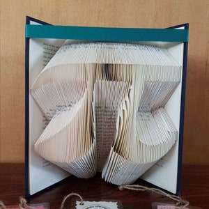 May include: A book with pages folded into the shape of the mathematical symbol pi. The book is bound in blue with a teal ribbon.