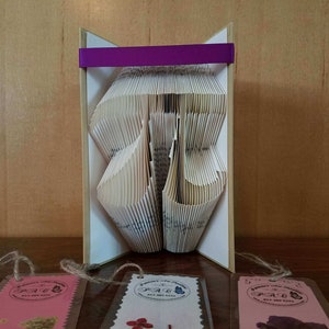May include: A handmade book sculpture with folded pages, forming a decorative shape. The sculpture is held within a light-colored frame and accented with a purple ribbon. Three small tags with floral designs and text are visible below.