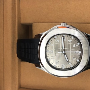 May include: A Seiko wristwatch featuring a silver-toned dial with a grid pattern, Arabic numerals, and luminous hands. The watch has a brushed stainless steel bezel and a black textured rubber strap with a secure buckle closure.