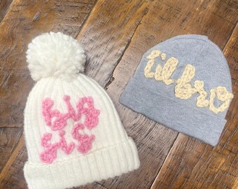 May include: Two knitted beanies. One is white with a pink "Big Sis" design and a white pom-pom. The other is light blue with a beige "Lil Bro" design. Both beanies are on a wooden surface.
