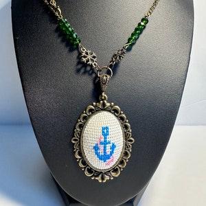 May include: A necklace with a green crystal and bronze chain. The pendant is an oval-shaped bronze frame with a white cross-stitch design of a blue anchor with pink flowers.