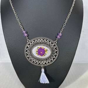May include: A silver chain necklace with a purple flower embroidered on a white fabric circle. The circle is surrounded by a silver filigree design and has a white tassel hanging from the bottom.