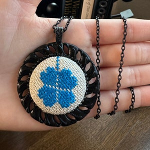 May include: A black chain necklace with a round pendant featuring a white fabric background with a blue embroidered flower design.