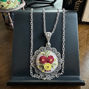 May include: A silver chain necklace with a round pendant featuring embroidered red and yellow flowers on a white fabric background. The pendant is surrounded by a silver filigree design.