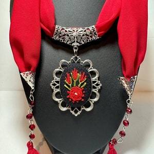 May include: A red silk scarf with a silver pendant featuring an embroidered floral design. The pendant is oval-shaped and has a black background with red and green embroidery. The scarf is tied around a black mannequin's neck.
