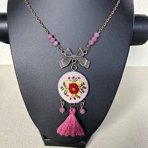 May include: A pink and white embroidered floral necklace with a pink tassel and a gold chain.