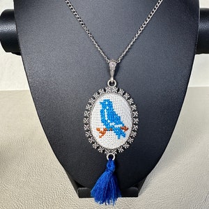 May include: A silver chain necklace with a blue bird cross-stitch design pendant in an oval frame. The pendant has a blue tassel.