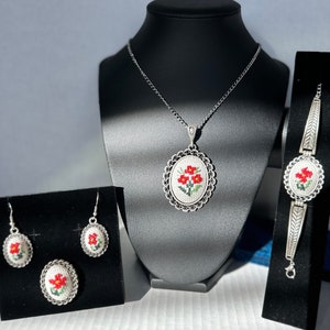 May include: A silver jewelry set with a necklace, bracelet, earrings, and a brooch. Each piece features a white oval with red embroidered flowers.