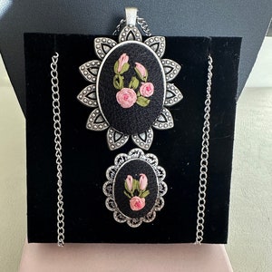 May include: A silver chain necklace with two black and silver pendants featuring pink embroidered flowers. The necklace is displayed in a box with the text "TAKE-N-GO TREASURE".