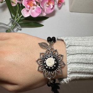 May include: A black and silver bracelet with a floral charm and black tassels. The charm has a white center with a black and white design.