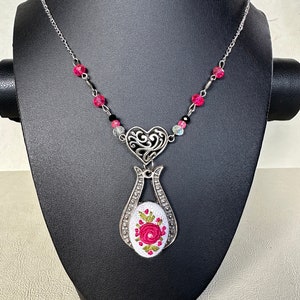 May include: A silver necklace with a heart-shaped pendant and a white fabric charm with pink embroidered flowers. The necklace is adorned with pink and black beads.