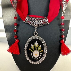 May include: A red scarf with a silver pendant featuring an embroidered white flower with green leaves on a black background. The pendant is attached to a silver chain with red beads and tassels.