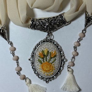 May include: A cream-colored scarf with a silver metal clasp and a pendant featuring an embroidered floral design in yellow and green. The pendant is surrounded by a silver metal frame. The scarf has two white tassels and a beaded chain with pink beads.