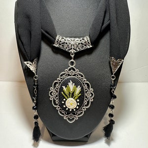 May include: A black choker necklace with a silver pendant featuring a white flower and green leaves embroidered on black fabric. The pendant is surrounded by an ornate silver frame. The necklace has two black tassel accents.