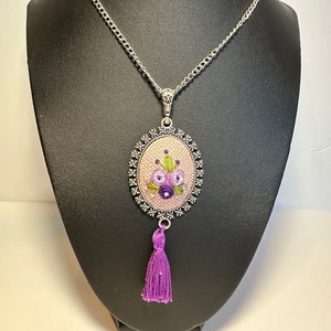 May include: A silver chain necklace with a purple and green embroidered floral pendant and a purple tassel.