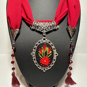 May include: A red fabric necklace with a silver pendant featuring an embroidered floral design. The pendant is oval-shaped and has a black background with red and green embroidery. The necklace has two tassel ends with red beads.