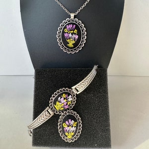 May include: A silver necklace and bracelet set with oval-shaped pendants featuring embroidered floral designs in shades of purple, yellow, and green on a black background.