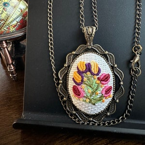 May include: A gold chain necklace with a pendant featuring a white fabric oval with embroidered flowers in shades of pink, purple, yellow, and green. The pendant is framed by a decorative metal border.