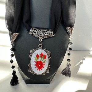 May include: A black silk scarf with a silver pendant featuring a white oval with red embroidered flowers. The pendant is attached to a silver chain with black beads and tassels.