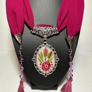 May include: A pink fabric necklace with a silver oval pendant featuring embroidered flowers. The pendant is attached to a silver chain with a silver clasp. The necklace has two pink tassels.