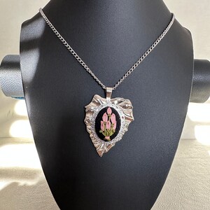 May include: A silver chain necklace with a pendant featuring a black fabric oval with pink embroidered flowers. The pendant is shaped like a leaf with a textured silver finish.
