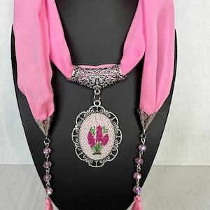 May include: A pink scarf with a silver pendant featuring a floral design. The pendant is attached to the scarf with a silver chain and has a beaded tassel on each end.
