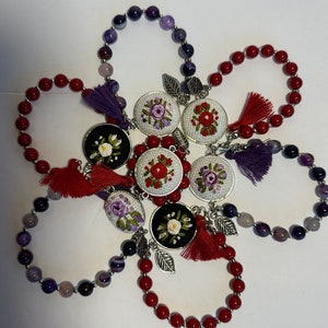 May include: Six beaded bracelets with embroidered floral charms. The bracelets are made with red, purple, and gray beads. The charms are silver with white and purple embroidery.