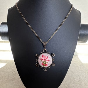 May include: A delicate necklace with a bronze chain and a round pendant featuring a pink embroidered floral design. The pendant is framed by a snowflake-shaped metal design.