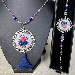 May include: A silver necklace with a blue tassel and a round pendant featuring a white embroidered floral design. The pendant is surrounded by a silver filigree frame. A matching silver bracelet with a similar floral pendant is also included.