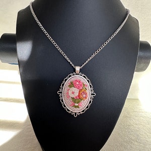 May include: A silver chain necklace with an oval pendant featuring a pink, white, and orange floral embroidery design.