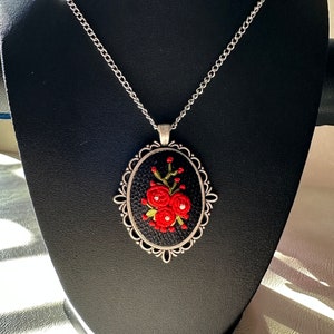 May include: A silver chain necklace with a black oval pendant featuring red embroidered flowers. The pendant has a silver ornate frame.