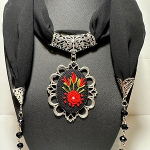 May include: A black scarf necklace with a silver pendant featuring an embroidered red flower design. The pendant is attached to a silver chain with black beads and tassels.