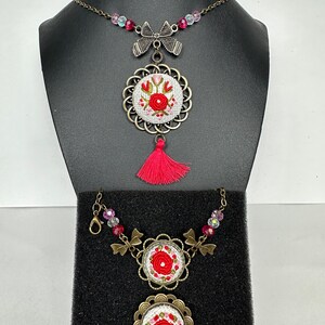 May include: A set of three necklaces with red and white embroidered floral designs. The necklaces feature a variety of beads and charms, including a bow, a tassel, and a floral motif.